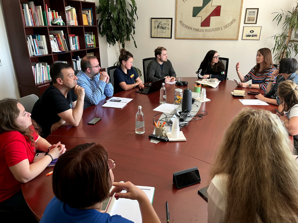 A group of project participants sit around an oval meeting table. One of the participants is speaking, the others are looking at her, apparently listening.