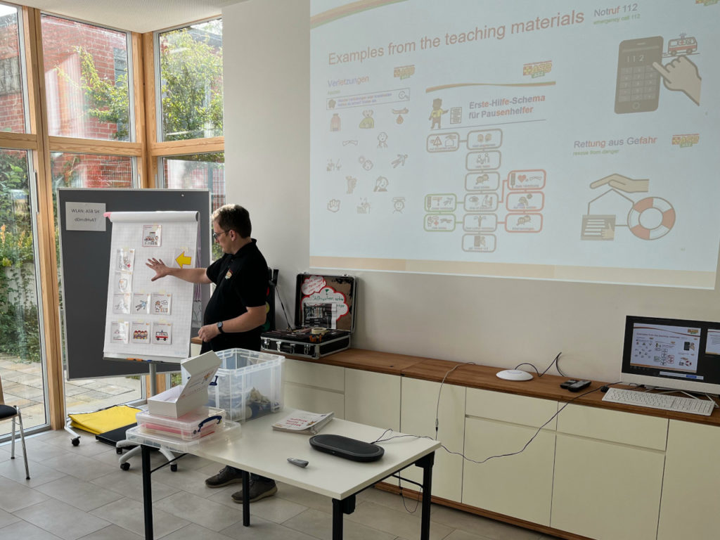 View of the front of a course room. Behind a teacher's table, Jens Vetter from ASB Schleswig-Holstein is seen explaining pieces of inclusive teaching materials pinned to a flip chart. Behind him, a presentation screen is showing more examples.